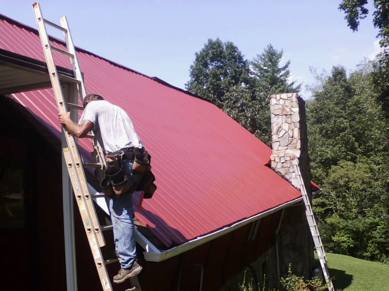 Roofer on a ladder beside a red metal roof and stone chimney — Soffit & Fascia Repair by Roofing Brothers in Mount Hermon